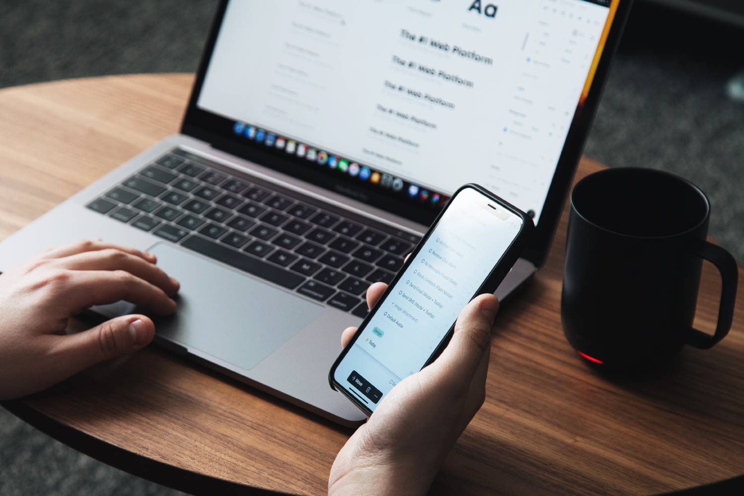 A person using a phone and laptop on a wooden table.